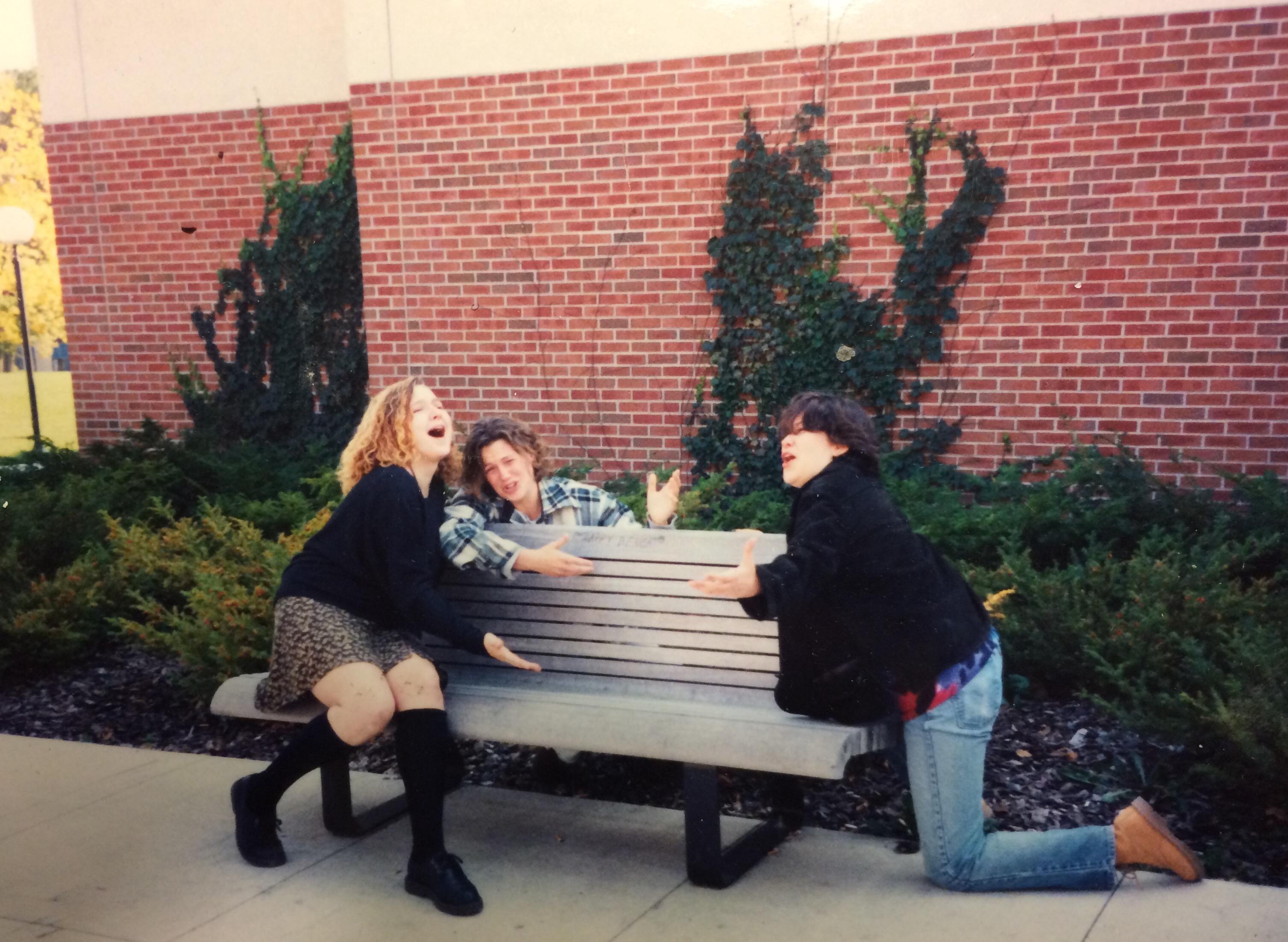 Three young women singing and gesturing as they sit on and around a bench near the Student Center on campus.