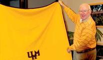 An older man holding up a gold blanket embroidered with "UMM."